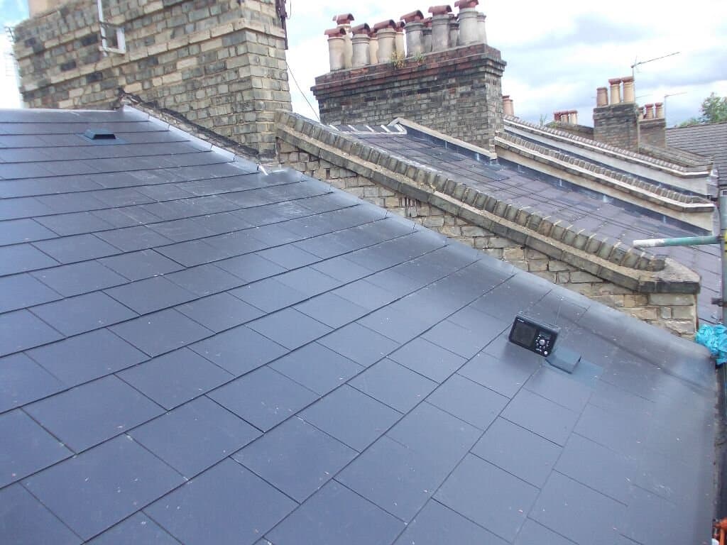 The image shows a close-up of a rooftops with slate tiles, chimneys, and roofing vents on a residential property. The sky is cloudy.